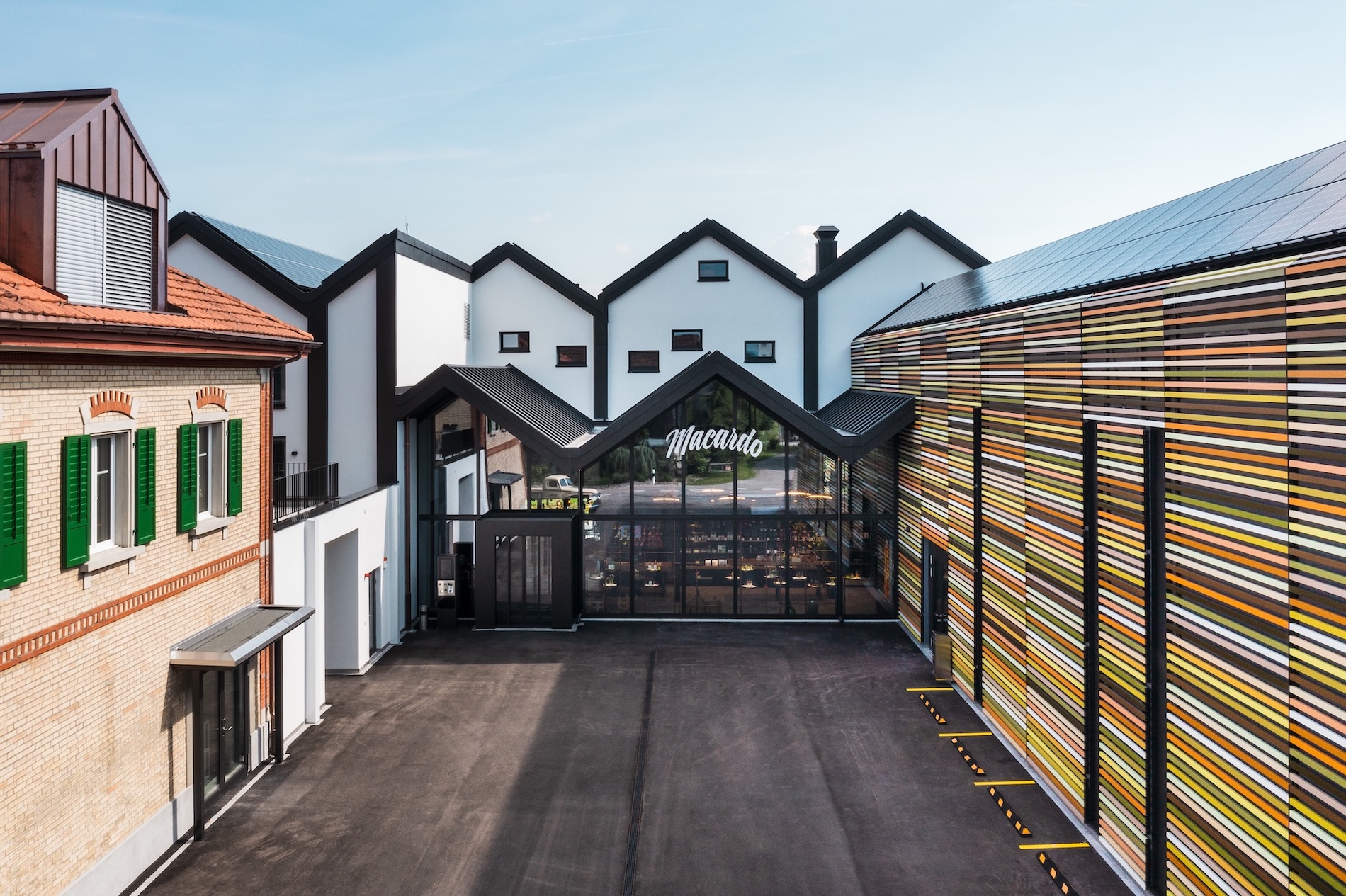 Übersicht über einen Hof mit mehreren Gebäuden, darunter eines mit horizontal gestreiften Paneelen in Gelb-, Braun- und Grüntönen. Das Hauptgebäude hat eine Glasfassade mit dem Schriftzug "Macardo". Weiße Giebel im Hintergrund.